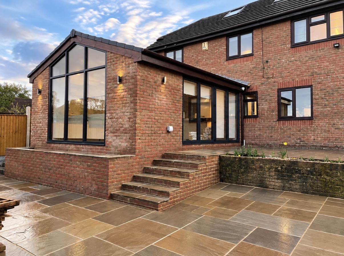Red-brick house with a single-storey rear extension, full-height black-framed gable window, open bi-fold doors and a sandstone paved patio with brick steps