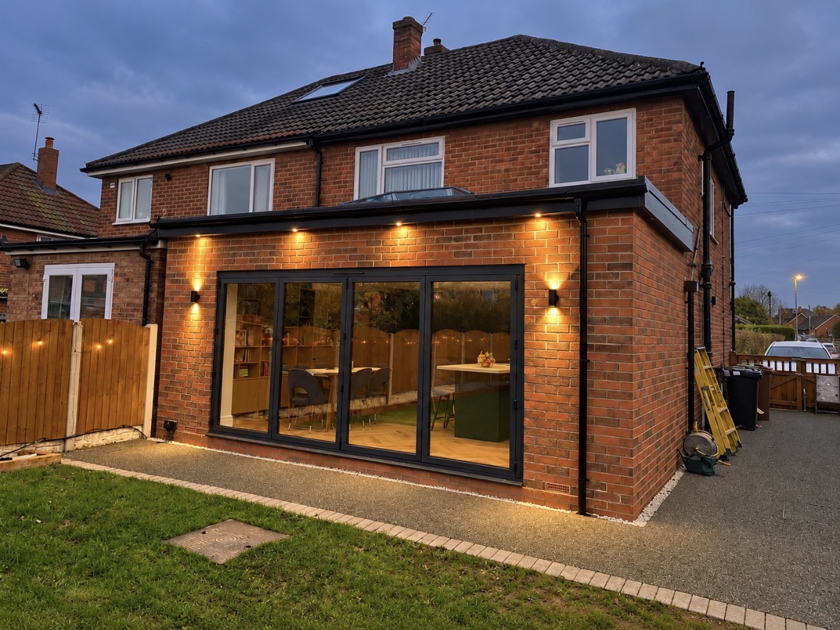Red-brick rear extension at dusk with a flat roof and glass roof lantern, anthracite grey bifold doors, lit soffit downlights and wall sconces, above a lawn with a paved path